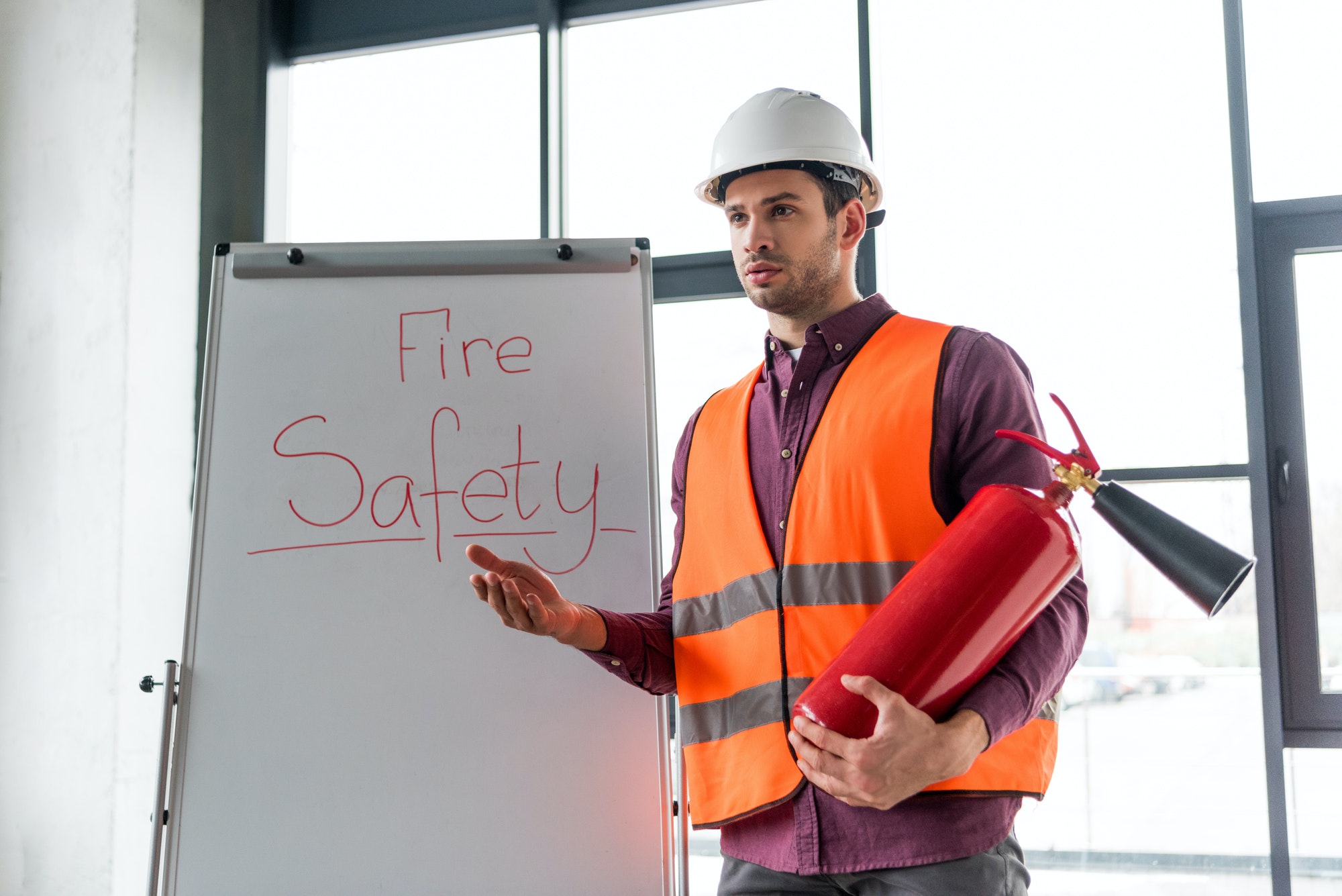 handsome fireman holding red extinguisher while standing near white board with fire safety lettering